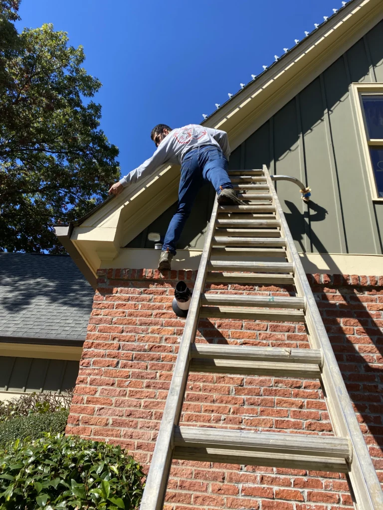 Installer working from a ladder along a residential roofline during a Christmas light installation.