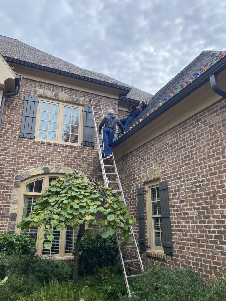 Installer on a ladder placing Christmas lights along the roofline of a brick residential home.