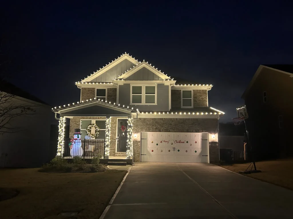 Residential home at night with warm white Christmas lights along rooflines, garage edges, and entry features.